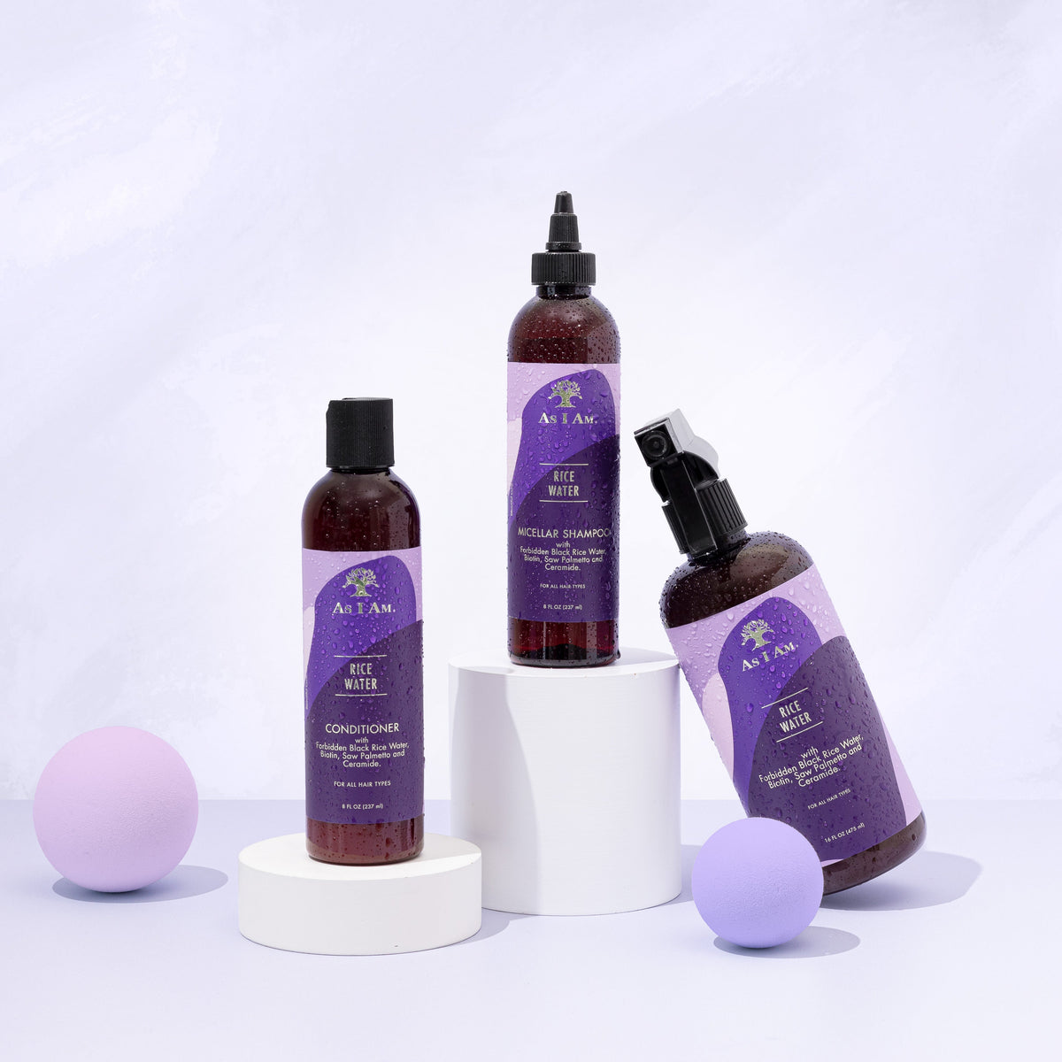 Tre produkter fra As I Am Rice Water-serien: Micellar Shampoo, Conditioner og Spray, er arrangeret på hvide podier med lilla kugler på en hvid baggrund.