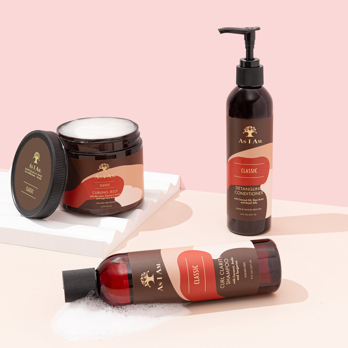Tre hårplejeprodukter fra As I Am står på en lys bordplade. Fra venstre mod højre ses en mindre flaske shampoo, en stor krukke med hårcreme og en høj flaske med balsam. Produkterne har alle en brun farve og en hvid etiket med gulddetaljer.