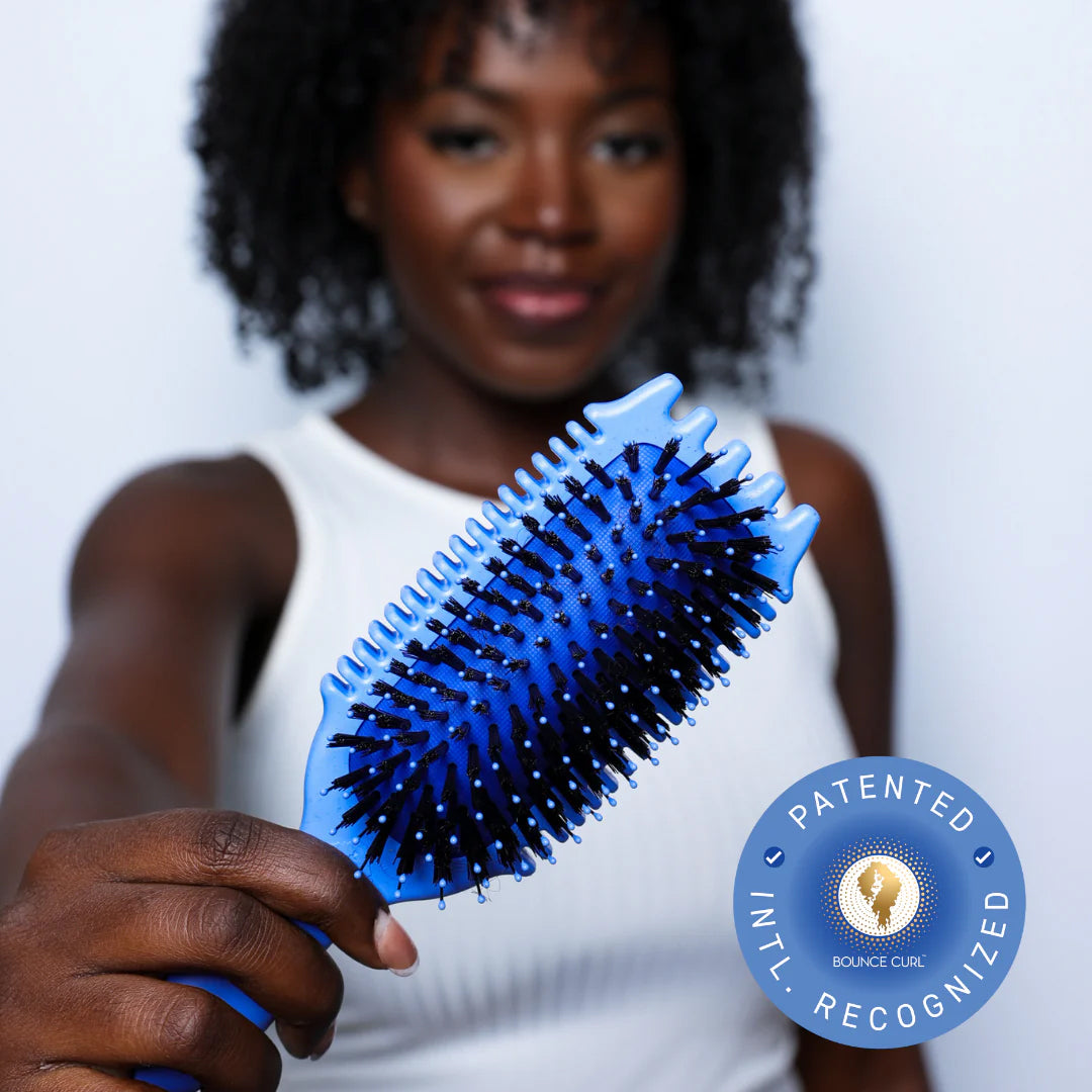 Kvinde med krøllet hår holder Bounce Curl Volume EdgeLift Brush Blue med blå og sorte børstepigge i fokus.