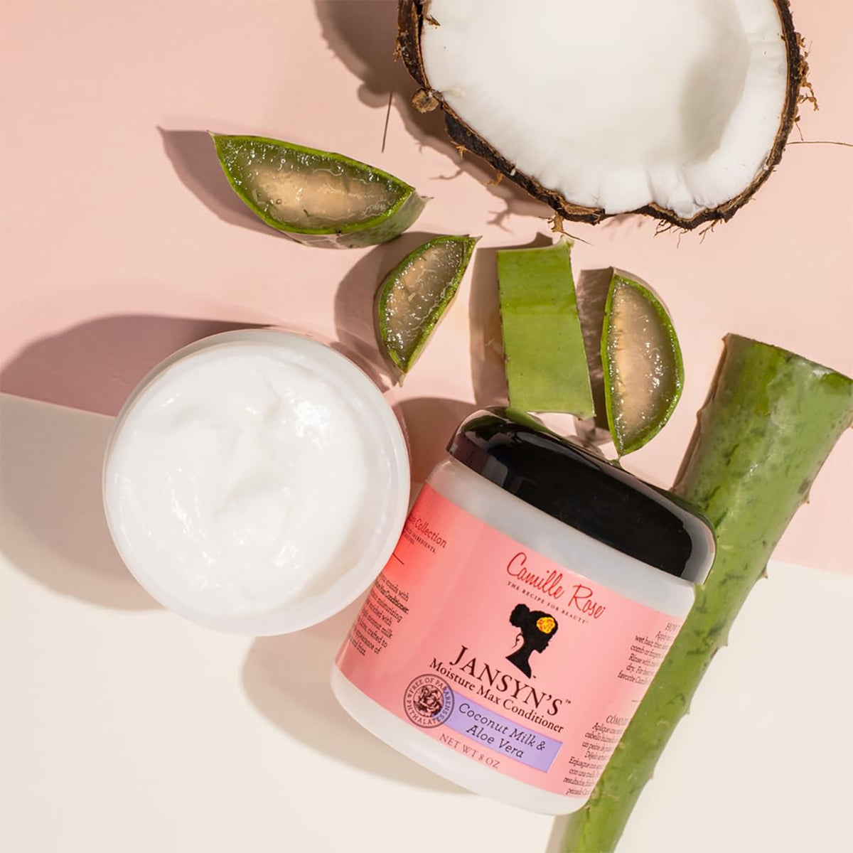 Camille Rose Jansyn's Moisture Max Conditioner i en hvid beholder med en sort top. Produktet er vist sammen med naturlige ingredienser som kokosnød og aloe vera.