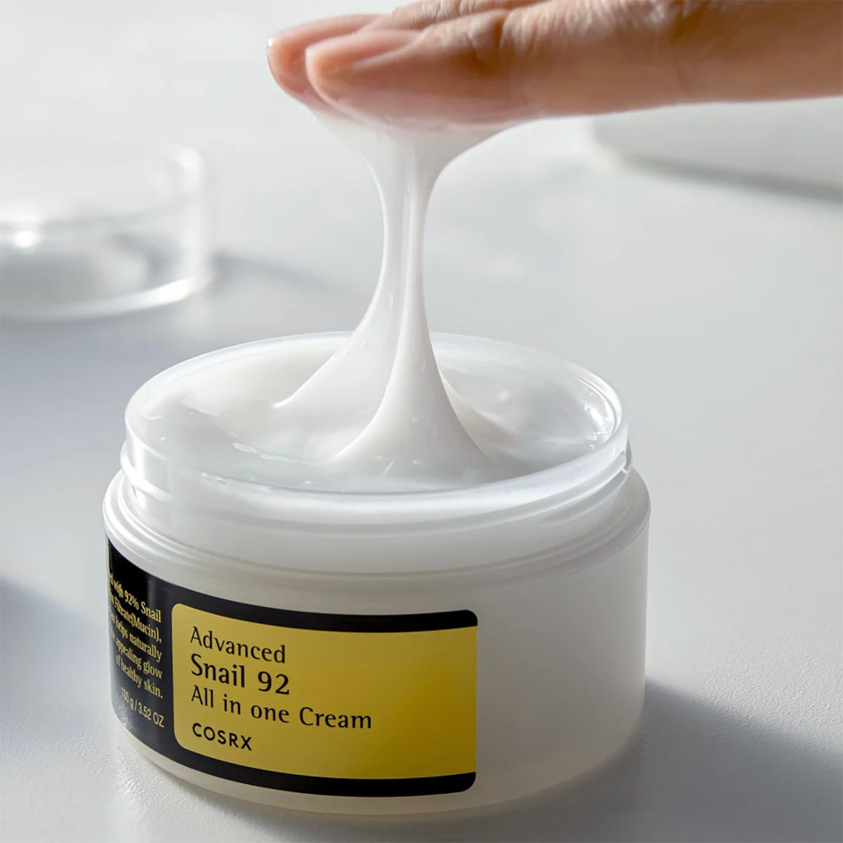 Fingre løfter elastisk tekstur fra åben krukke med Cosrx Advanced Snail 92 Cream 100gr på hvidt bord.