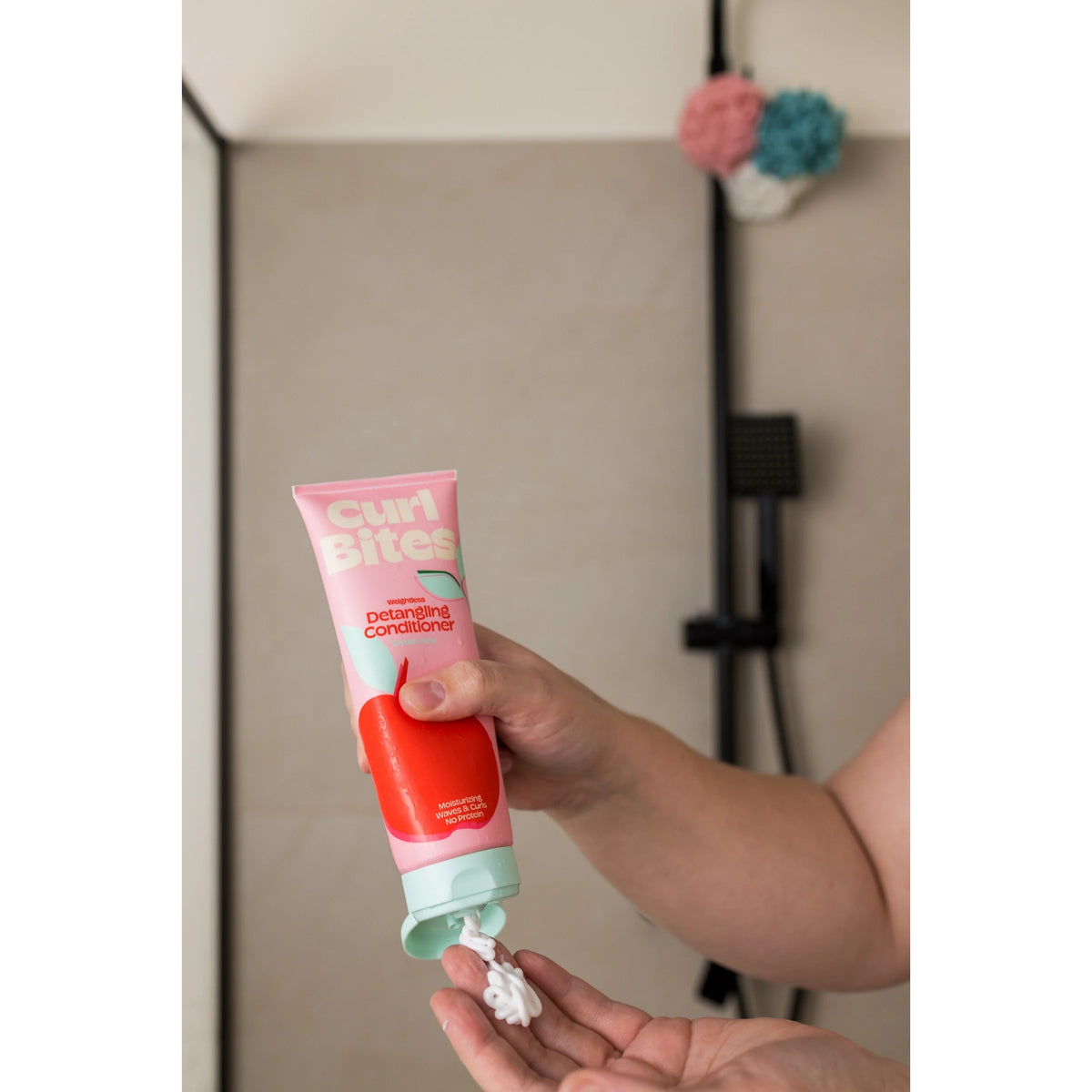 Hånd klemmer Curl Bites Detangling Conditioner ud i pink tube med æblemotiv i badeværelse.