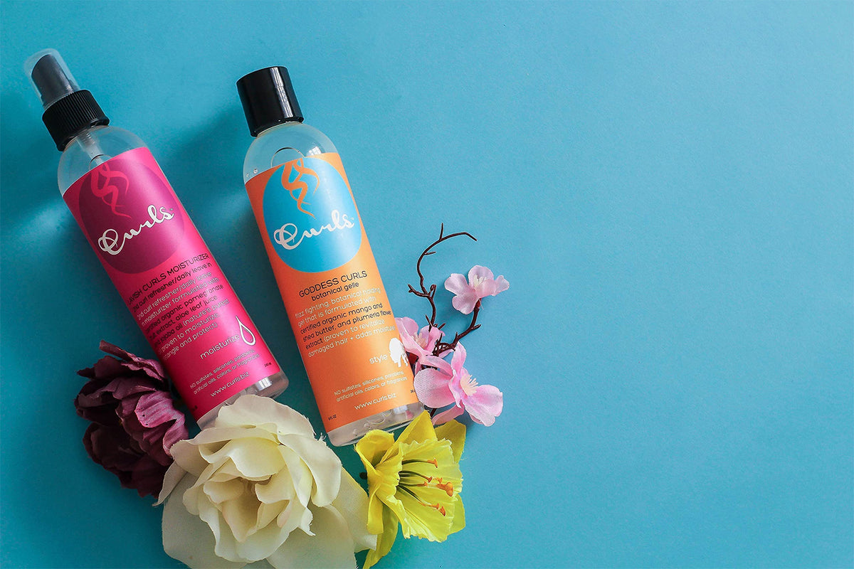 Curls Lavish Curls Moisturizer (pink) og Goddess Curls Botanical Gelle (orange) på blå baggrund med blomster. Hårplejeprodukter til krøller.