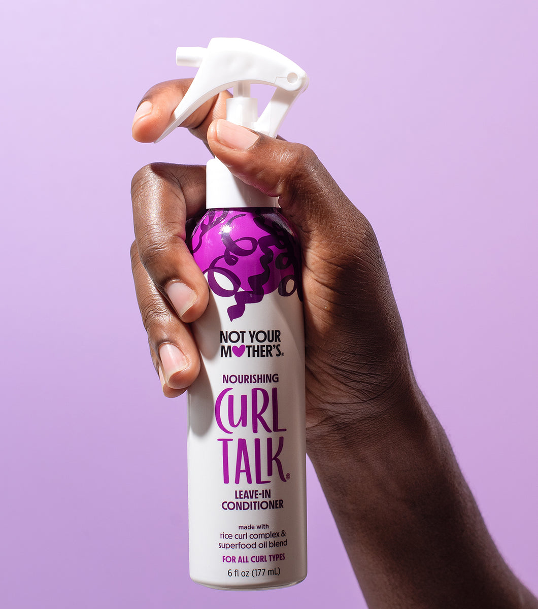 En mørk hånd sprayer Not Your Mother's Curl Talk Nourishing Leave-In Conditioner foran en ensfarvet lilla baggrund.