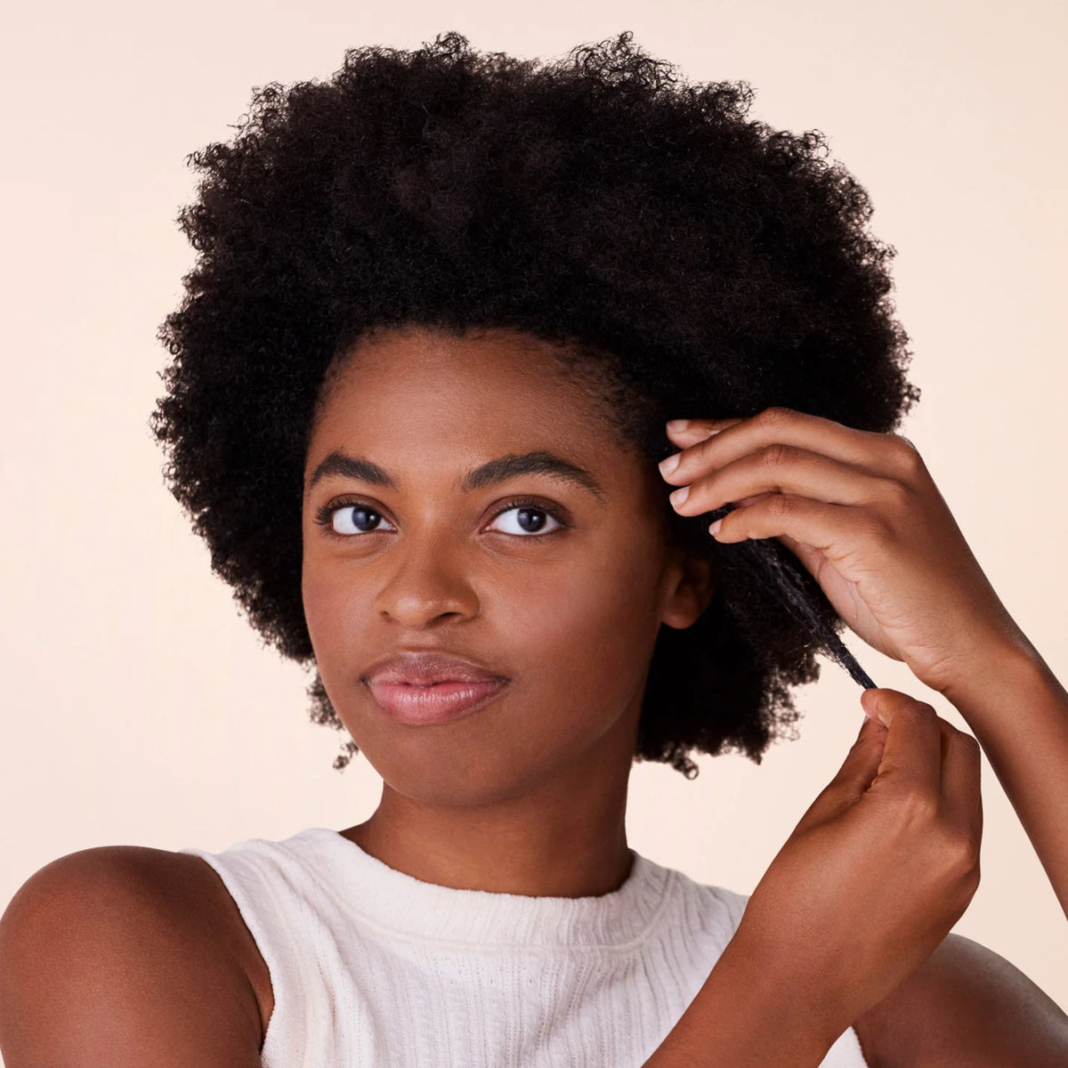 Kvinde med naturligt afrohår sektionerer sit krøllede hår med fingrene mod en beige baggrund.