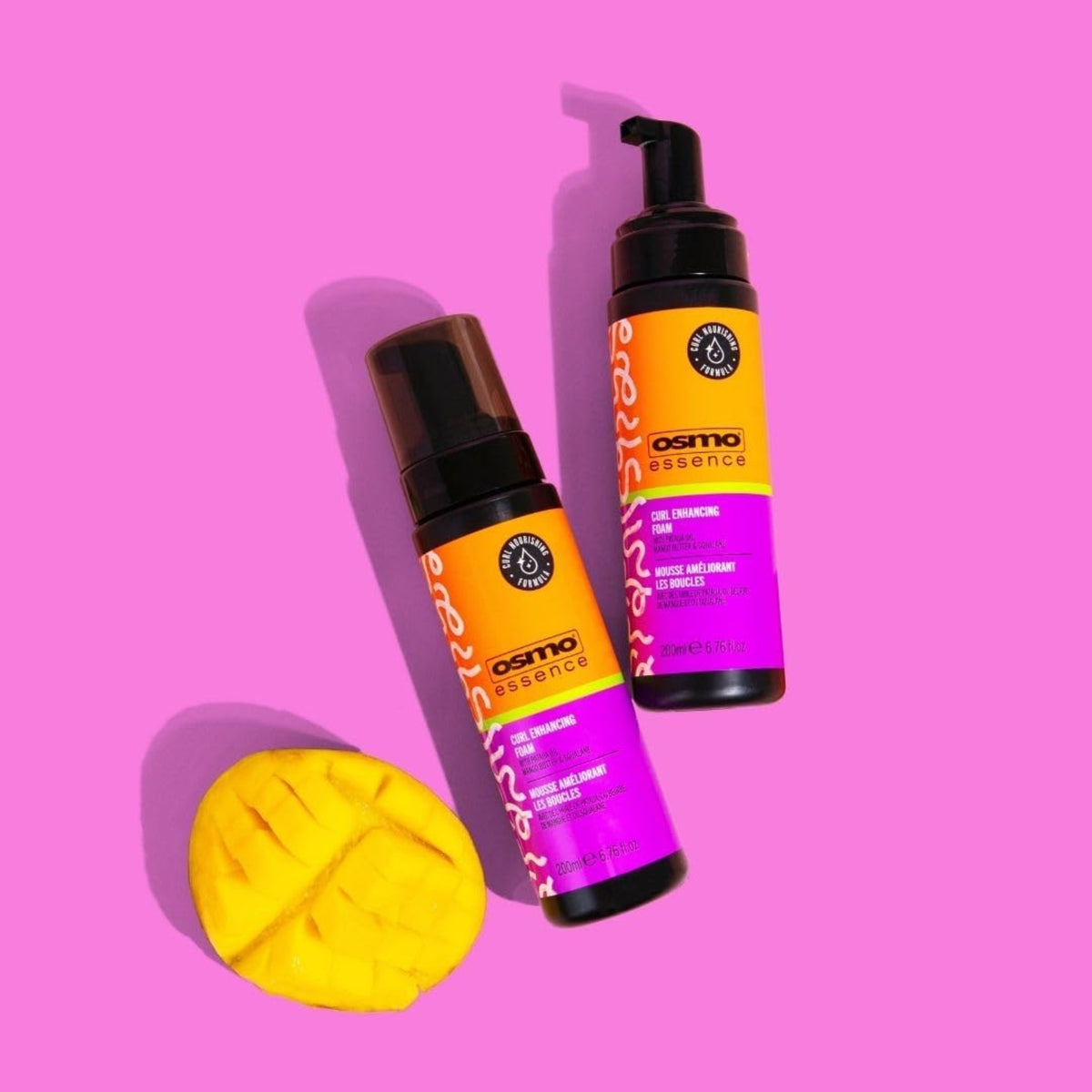 To flasker Osmo Essence Curl Enhancing Foam i orange/lilla emballage ved siden af en udskåret mango på pink baggrund.