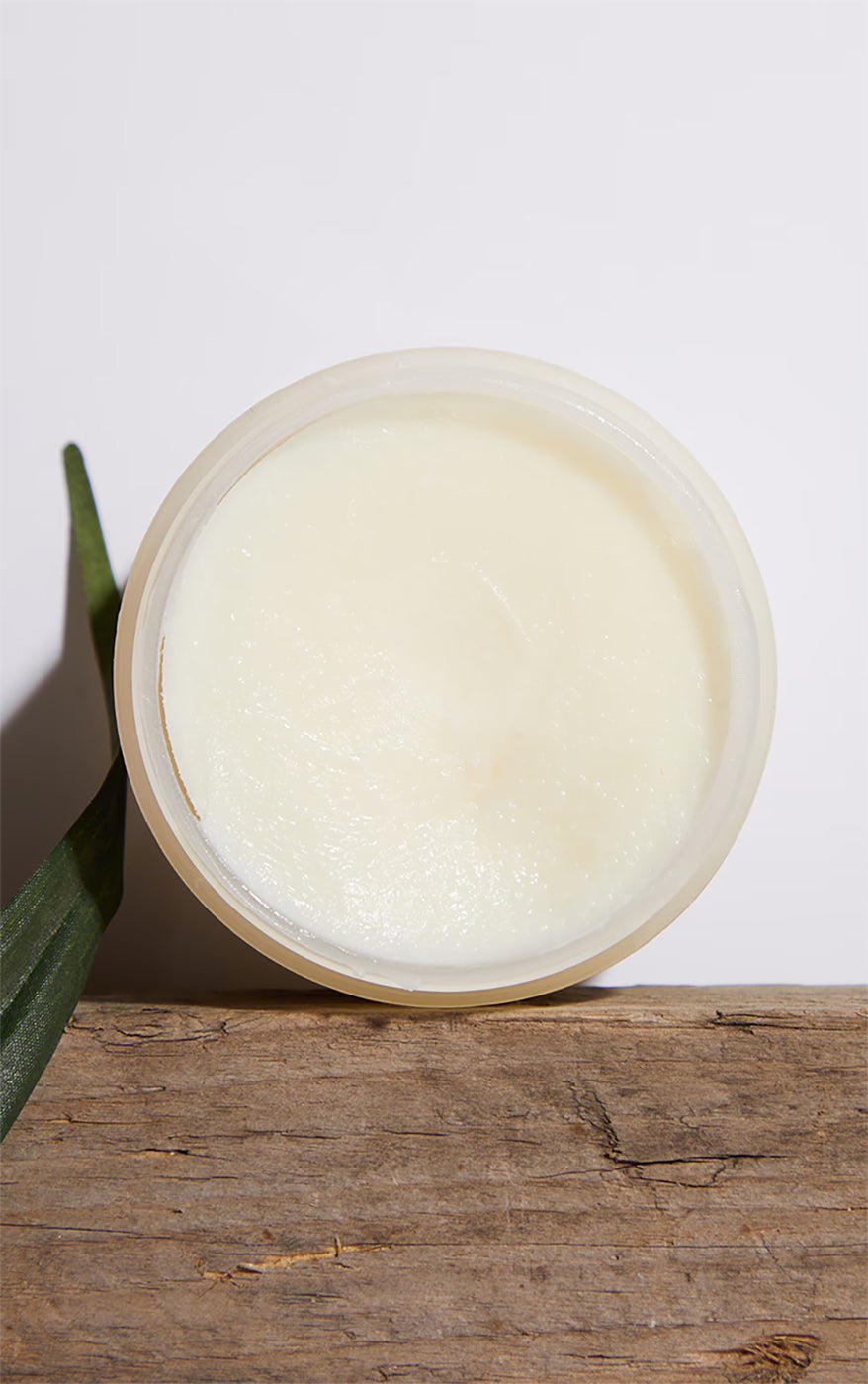 Palmers Coconut Oil Formula Body Balm i åbent på rustik træoverflade med grønt blad.
