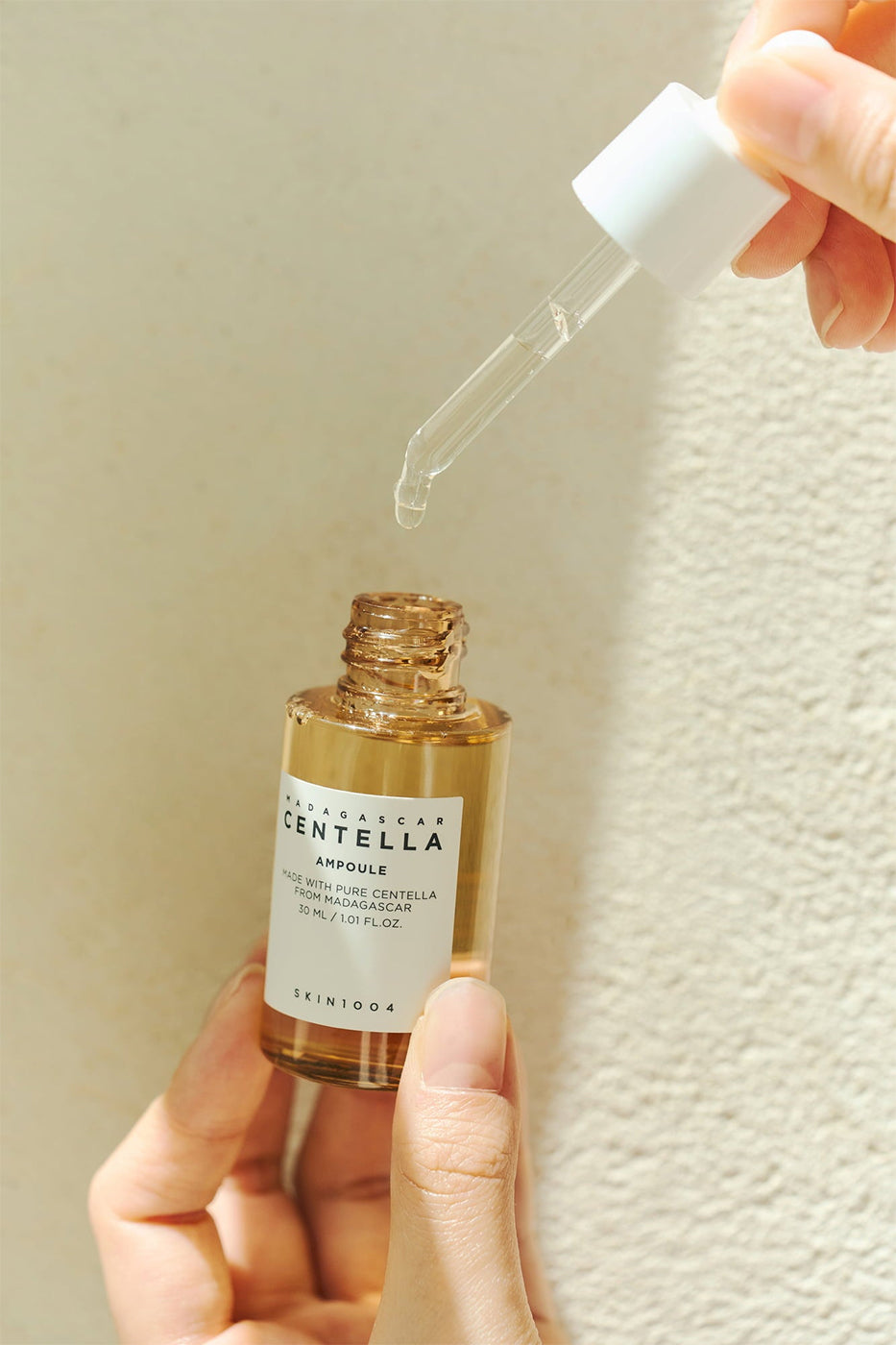 Pipette med serum fra SKIN1004 Madagascar Centella Ampoule 100ml løftes over gylden flaske i sollys mod lys væg.