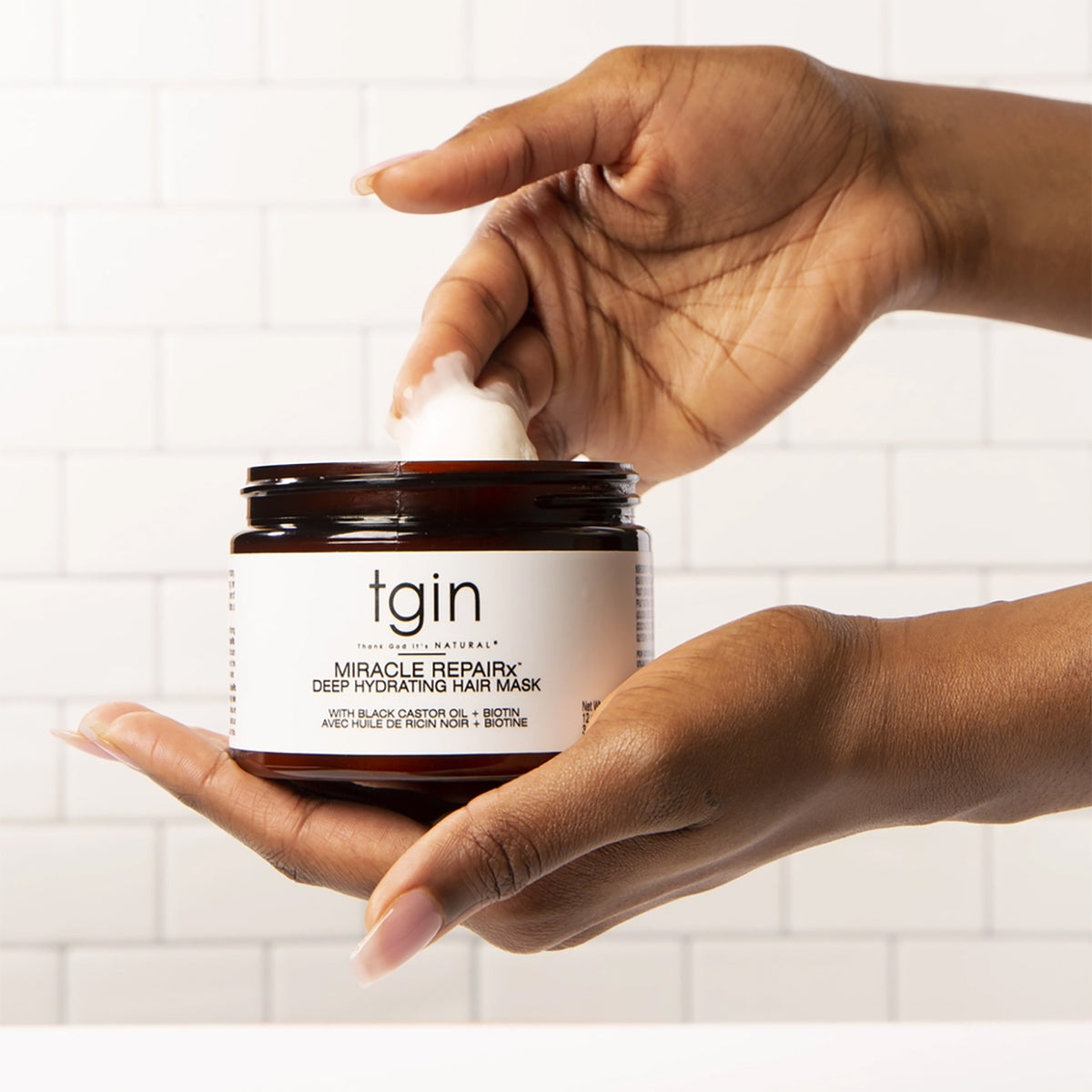 Hånd dypper fingre i krukke med TGIN Miracle RepairX Deep Hydrating Mask 340gr foran hvid flisevæg.
