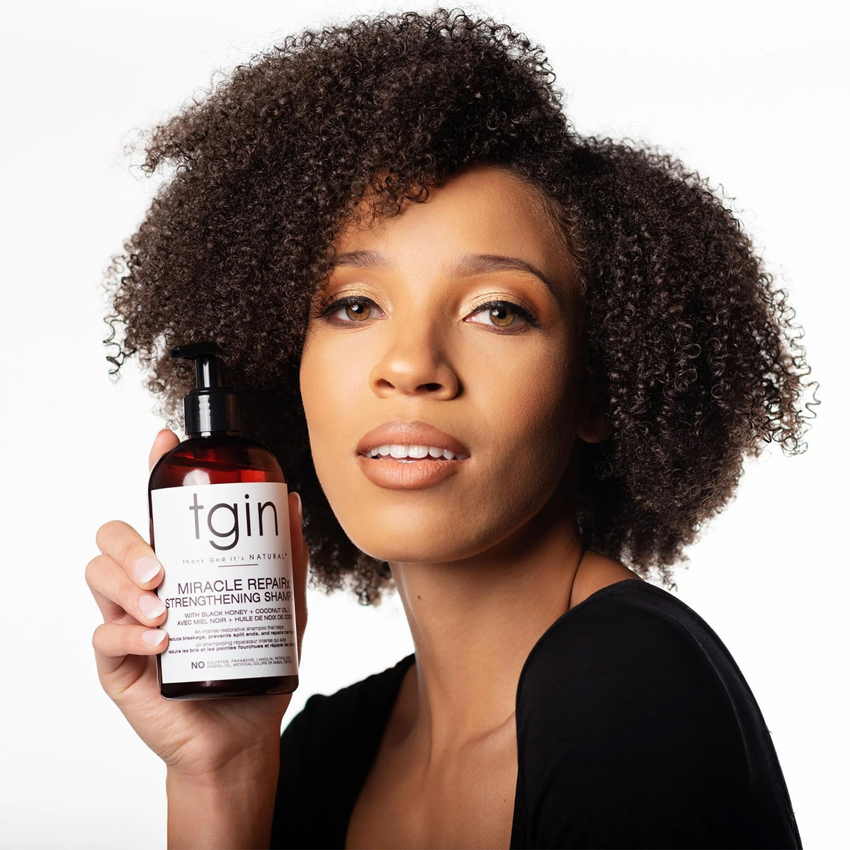 Kvinde med krøllet hår holder TGIN Miracle RepairX Strengthening Shampoo 384ml foran ansigtet mod hvid baggrund.